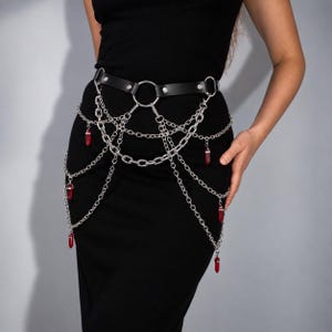 May include: A black leather belt featuring a silver-toned metal ring at the centre, adorned with multiple layered silver chains that drape downwards. Several small, pointed, deep red crystal-like pendants hang from the ends of the silver chains.