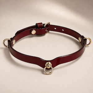 Op de afbeelding: Een dieprode, glanzende chokerhalsband met goudkleurige metalen hardware. Het ontwerp bevat een centrale O-ring, twee zijdelingse O-ringen en een verstelbare gespsluiting met een bijpassende lus, alles vastgezet met goudkleurige klinknagels.