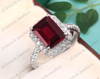 May include: A silver ring featuring a large, rectangular, deep red gemstone surrounded by a halo of smaller, clear stones. The ring's band is also adorned with small, clear stones, creating a sparkling effect. The ring is set against a soft, neutral background.