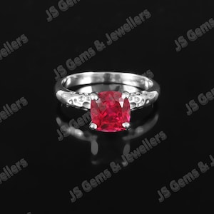 Peut inclure: Une bague en argent ornée d'une grosse pierre rouge vif de forme carrée. Le corps de la bague présente un motif texturé. La bague est présentée sur un fond noir. Les mots "JS Gems & Jewellers" sont visibles.