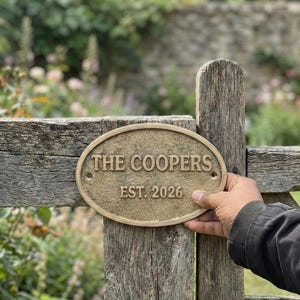 May include: An oval, bronze-colored plaque with the text "THE COOPERS EST. 2026" mounted on a weathered wooden fence. The plaque is held by a person's hand, with a blurred garden background.