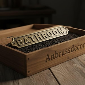 May include: A brass "BATHROOM" sign rests in a wooden box filled with dark granules. The sign is rectangular with rounded edges. The box is engraved with "Anbrassdecor" on the side, and sits on a wooden surface.