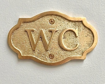May include: A gold-colored metal sign with a textured surface and a smooth border. The sign features the letters WC in a raised, serif font. Two small screws are visible at the top and bottom center of the plaque, securing it to the wall.