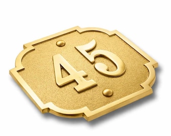 May include: A gold-colored metal house number plaque featuring the digits 45 in a raised, polished finish against a textured, matte background. The plaque has a decorative, scalloped-edge shape with two small, circular mounting points.