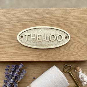 May include: An oval, antique brass sign with the text "THE LOO" is mounted on a light brown wooden surface. The sign is surrounded by a decorative border. Below the sign are a rolled white cloth, a vintage key, and a small glass bottle.