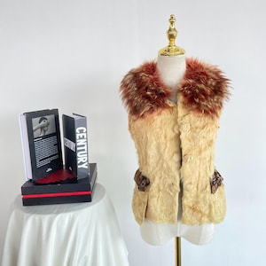 Puede incluir: Un chaleco de piel de color beige con cuello de piel marrón y rojo. El chaleco tiene dos bolsillos decorativos con un estampado marrón y beige y está abotonado en la parte delantera. El chaleco se exhibe en un maniquí.
