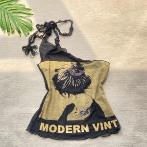 Puede incluir: Top halter negro y dorado con un diseño decorativo y el texto "MODERN VINT". El top presenta una silueta única y un gráfico de inspiración vintage. Fabricado con un material ligero.