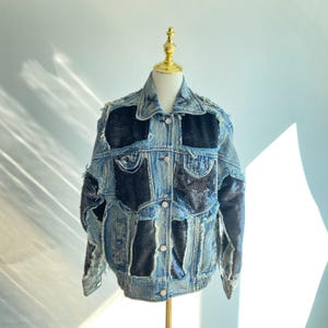 Puede incluir: Chaqueta vaquera azul claro con parches de lentejuelas negras y detalles desgastados. La chaqueta presenta un cuello clásico, cierre de botones en la parte delantera y mangas largas. La chaqueta se exhibe en un maniquí dorado.