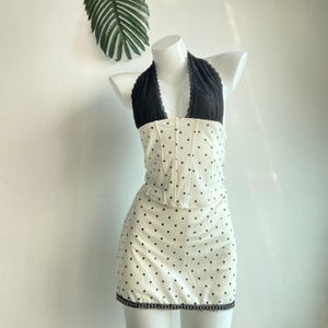 Puede incluir: Conjunto de dos piezas sobre un maniquí. La parte superior es un top halter negro con ribete de encaje, combinado con un corpiño blanco estilo corsé con lunares negros. La falda a juego es blanca con lunares negros y ribete de encaje negro.