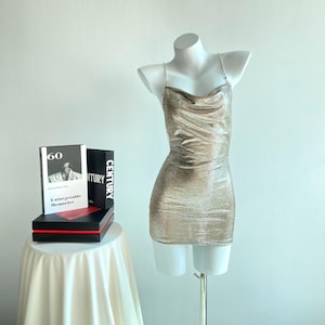 Puede incluir: Un minivestido dorado y brillante con tirantes finos expuesto en un maniquí blanco. El vestido tiene un cuello drapeado y un corte ajustado. Dos libros están sobre una mesita a la izquierda del maniquí.