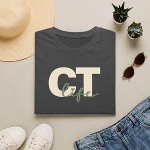 Könnte beinhalten: Ein dunkelgraues T-Shirt mit dem Text CT in großen cremefarbenen Blockbuchstaben und dem Wort life in salbeigrüner Schreibschrift darunter, inklusive eines kleinen Herzsymbols über dem Buchstaben i im Wort life.