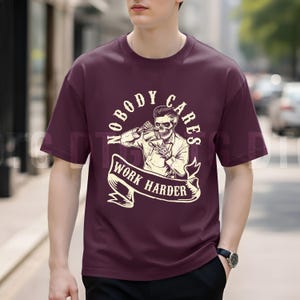 May include: Burgundy crew neck t-shirt featuring a graphic of a skeleton pouring a drink, with the text "NOBODY CARES WORK HARDER" in a banner. The design is in cream and black.