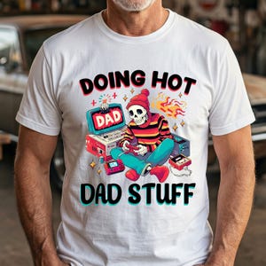 Può includere: T-shirt bianca con una grafica colorata di uno scheletro con berretto rosso e camicia a righe, che gioca a un videogioco. Il testo "DOING HOT DAD DAD STUFF" è stampato sopra e sotto la grafica.