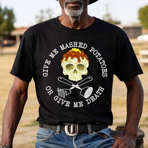 Puede incluir: Una camiseta negra con un gráfico de una calavera cubierta de puré de patatas y un trozo de mantequilla. Debajo de la calavera hay dos pasapurés cruzados. El texto que rodea el diseño dice, GIVE ME MASHED POTATOES OR GIVE ME DEATH.