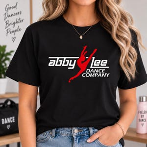 May include: A black short-sleeved t-shirt featuring a white graphic logo that reads "abby lee DANCE COMPANY" with a red silhouette of a dancer in a leap pose integrated into the text. The shirt has a crew neckline and a standard fit.