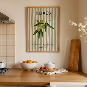 May include: A framed print with the word "OLIVES" in black, and an olive branch illustration. The background has green and white stripes. The text "handpicked produce, est. 1973" is below the olive branch. The print is in a kitchen.