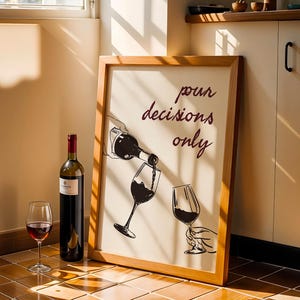 May include: A framed artwork with the text "pour decisions only" next to a wine bottle and a glass of red wine. The artwork depicts wine being poured into a glass. A full bottle of red wine and an empty wine glass are also present.