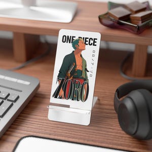 May include: A white phone stand featuring a graphic of the character Zoro from the anime series One Piece. The image shows Zoro in a green jacket and the text "ONE PIECE" and "ZORO".