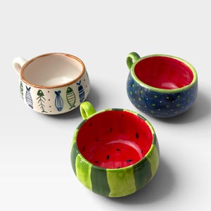 Pode incluir: Três canecas de cerâmica com designs distintos. Uma apresenta um exterior branco com padrões de peixes azuis e verdes. Outra é azul-escura com bolinhas brancas e interior vermelho. A terceira assemelha-se a uma melancia com riscas verdes.