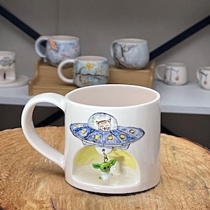 May include: White ceramic mug with a whimsical design featuring a cat in a blue UFO, with a green alien figure hanging below. The mug has a large handle and is surrounded by other decorative mugs on a wooden surface.