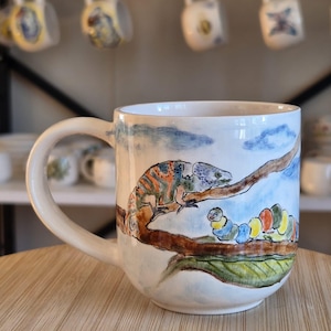 May include: A white ceramic mug featuring a hand-painted illustration of a chameleon perched on a brown tree branch, with a colorful segmented caterpillar crawling below it on a large green leaf against a light blue sky with clouds.