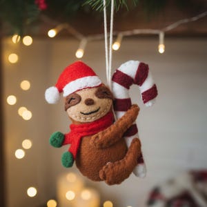 May include: A handmade Christmas ornament featuring a brown sloth wearing a red Santa hat, scarf, and green mittens. The sloth is hugging a red and white striped candy cane. The ornament is suspended by a white string.