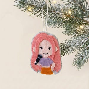 May include: A felt Christmas tree decoration depicting a cartoon figure with pink hair, a purple top, and an orange skirt, holding a black microphone. The ornament hangs from a white string against a snowy green branch.
