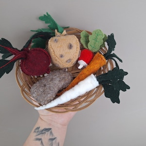 Könnte beinhalten: Ein Weidenkorb gefüllt mit Filzgemüse, darunter eine rote Bete, eine Karotte und ein Pilz. Das Gemüse hat verschiedene Farben, darunter Rot, Orange und Grün. Der Korb wird von einer Person gehalten.