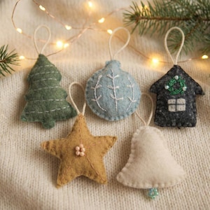 Può includere: Una collezione di ornamenti natalizi fatti a mano in feltro. Gli ornamenti includono un albero verde, una palla blu, una casa nera, una stella dorata e una campana bianca. Ogni ornamento ha un anello per appenderlo ed è decorato con cuciture e piccoli dettagli.