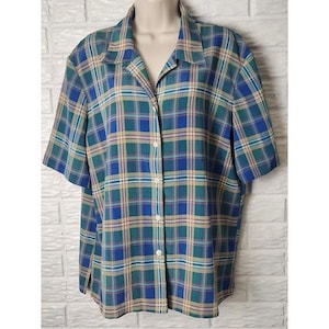 May include: A short-sleeved button-down shirt in a plaid pattern. The shirt features a color palette of blue, green, and tan. The shirt is displayed on a mannequin against a white brick wall.
