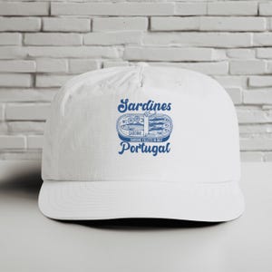 May include: White baseball cap featuring a blue graphic of a sardine can with the words "Sardines" and "Portugal". The text "Sardine Fillets in Oily" is also included. The cap has a curved peak.