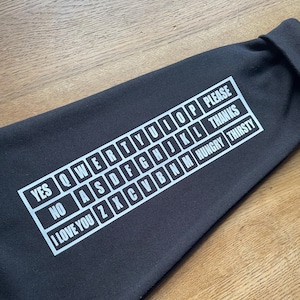 May include: A black long-sleeve garment featuring a white graphic print of a communication board. The print includes a QWERTY keyboard layout alongside words such as YES, NO, I LOVE YOU, PLEASE, THANKS, HUNGRY, and THIRSTY arranged in a grid.