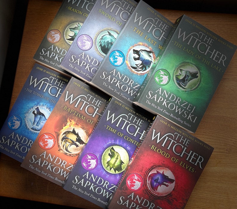 Op de afbeelding: Een verzameling van negen paperback boeken met kleurrijke omslagen met een wolfontwerp en de titel "The Witcher" in witte tekst. De boeken zijn van auteur Andrzej Sapkowski en maken deel uit van een New York Times bestseller-serie.