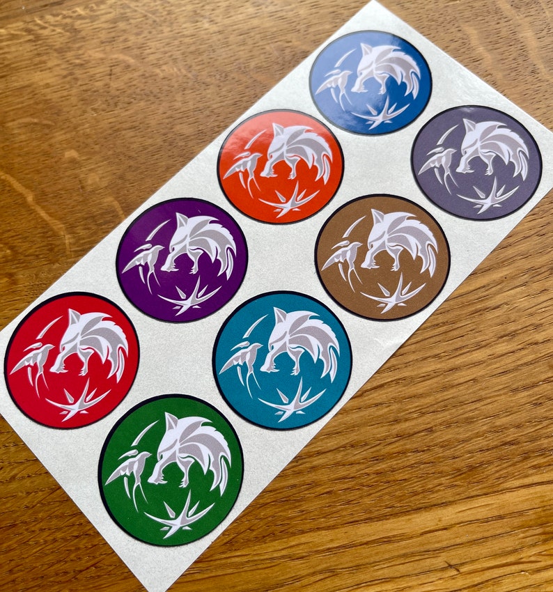 Op de afbeelding: Acht ronde stickers met een wit wolfskopontwerp met een vogel aan elke kant. De stickers zijn in verschillende kleuren, waaronder blauw, oranje, paars, bruin, rood, teal en groen.