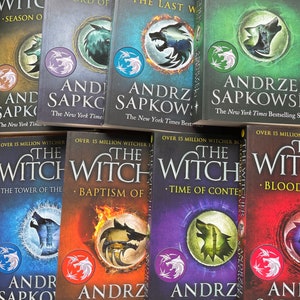 Op de afbeelding: Een verzameling van acht paperback boeken met kleurrijke omslagen met een wolf-afbeelding en de tekst "The Witcher" en "Andrzej Sapkowski".