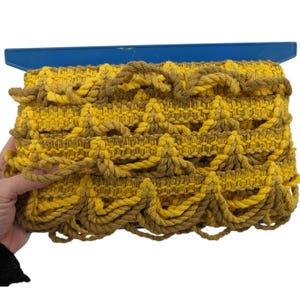 May include: A decorative trim featuring a repeating scalloped pattern in alternating shades of bright yellow and muted brown yarn. The textile is mounted on a flat blue plastic support, showcasing a textured, woven design with looped edges.