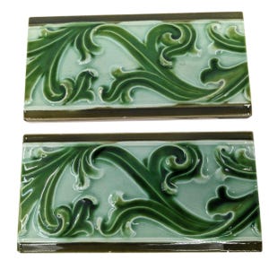 May include: Two rectangular green ceramic tiles with a repeating floral design. The tiles have a glossy finish and a dark green border. The floral pattern is a lighter shade of green and is raised from the surface.