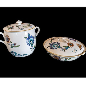 May include: A white ceramic lidded tureen and a matching shallow lidded serving dish, both decorated with a repeating floral pattern in shades of blue, orange, and green, featuring gold-toned trim along the edges and handles.