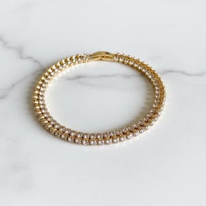 Puede incluir: Una pulsera dorada con dos filas de pequeños cristales transparentes. La pulsera es circular y tiene un cierre de broche. Los cristales brillan sobre el engaste dorado, creando un aspecto glamuroso. La pulsera se exhibe sobre una superficie de mármol blanco.