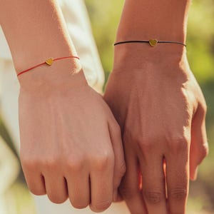 May include: Two hands holding each other, each wearing a thin string bracelet with a small gold heart charm. One bracelet is red, the other is black. The background is blurred green.