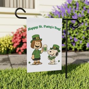May include: A white garden flag with the text "Happy St. Patty's Day!" in green. The flag features a cartoon girl and Snoopy, both wearing green hats and holding a beer and shamrocks. The flag is displayed on a black metal stand.