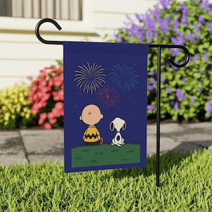 Może przedstawiać: Granatowa flaga ogrodowa przedstawiająca Charliego Browna i Snoopy'ego oglądających fajerwerki. Flaga ma ciemnoniebieskie tło z żółtymi, czerwonymi i niebieskimi fajerwerkami. Charlie Brown ma na sobie żółtą koszulę, a Snoopy jest biały z czarnymi plamami.