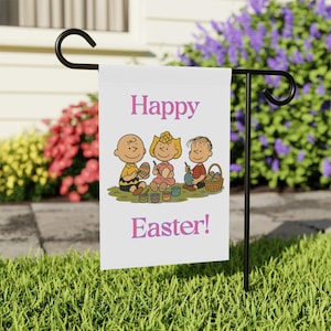 Może przedstawiać: Biała flaga ogrodowa z różowymi słowami "Happy Easter!". Flaga przedstawia kreskówkową ilustrację trojga dzieci malujących pisanki. Flaga jest wyświetlana na czarnym metalowym stojaku w ogrodzie.