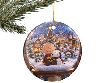 May include: A round ceramic Christmas ornament featuring a printed scene of Charlie Brown and Snoopy sitting in the snow. The background shows a festive town with a lit Christmas tree, a bridge, and a sign reading Christmas Town.