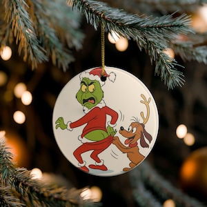 May include: A round white ceramic ornament featuring a cartoon illustration of a green character in a red Santa suit with a white trim, looking annoyed while a brown dog with antlers pulls down the back of the character's red pants.
