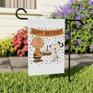 Może przedstawiać: Biała flaga ogrodowa z kreskówkową ilustracją Charliego Browna i Snoopy'ego. Charlie Brown trzyma tort urodzinowy ze świeczką, Snoopy stoi obok. Na banerze napis "HAPPY BIRTHDAY!" z konfetti. Flaga jest na czarnym metalowym stojaku.