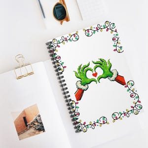 May include: A spiral-bound notebook featuring a white cover with a graphic of two green furry hands forming a heart shape around a small red heart, surrounded by a decorative border of colorful Christmas lights on black wire strings.