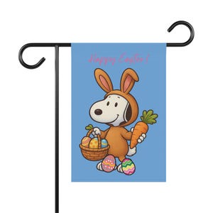 Może przedstawiać: Niebieska flaga ogrodowa z Snoopy'm w brązowym kostiumie królika, trzymającym marchewkę i kosz jajek wielkanocnych. Flaga ma różowy napis "Happy Easter!".