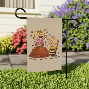 Pode incluir: Uma bandeira de jardim retangular bege pendurada num suporte de metal preto. A bandeira apresenta uma ilustração de desenho animado de uma menina loira com um vestido rosa a saltar para uma pilha de folhas de outono castanhas ao lado de um rapaz.