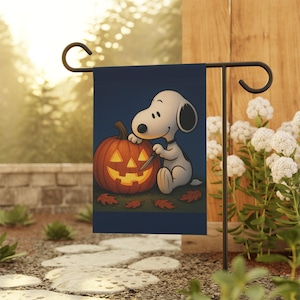 May include: A rectangular garden flag featuring a cartoon dog with white fur and black ears sitting next to a glowing orange jack-o'-lantern. The dog holds a small carving tool. Several brown autumn leaves are scattered on the ground below.